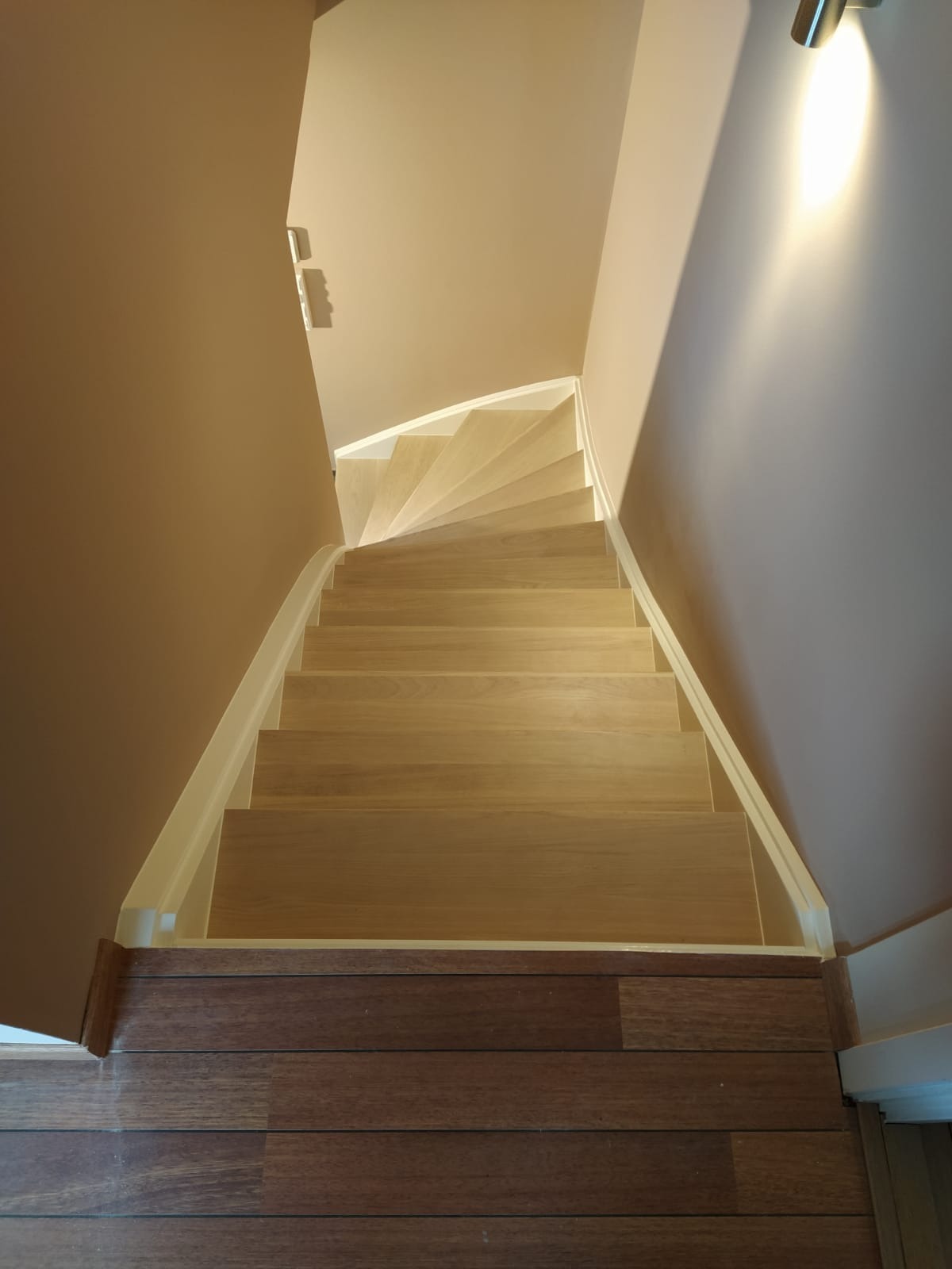 Jasne, drewniane schody z białymi listwami przypodłogowymi prowadzące w górę, widok z góry na tle beżowych ścian i podłogi z ciemnego drewna.