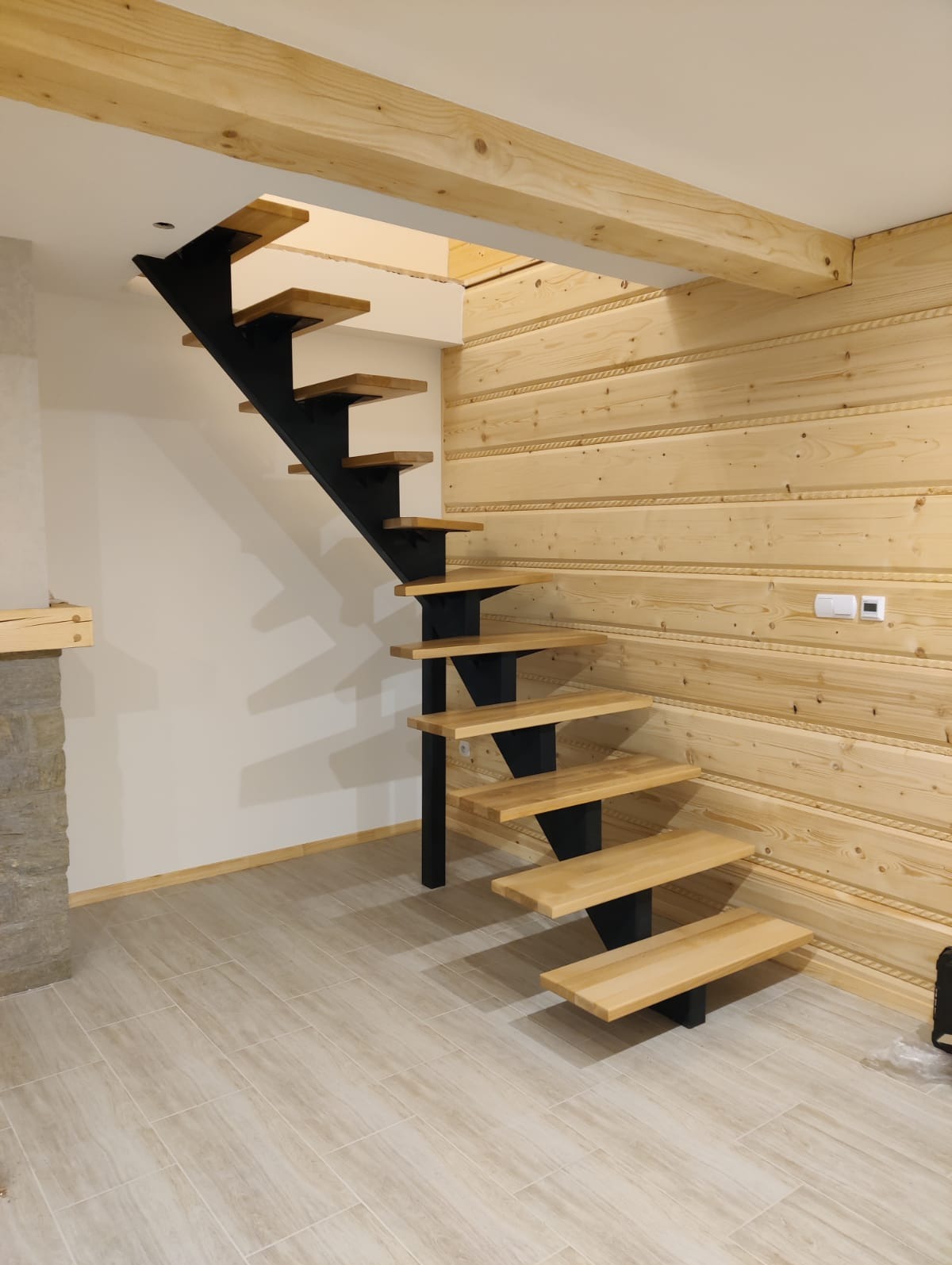 Nowoczesne schody drewniane z czarnymi metalowymi wspornikami, zamontowane w pomieszczeniu z boazerią i płytkami podłogowymi w odcieniach beżu.