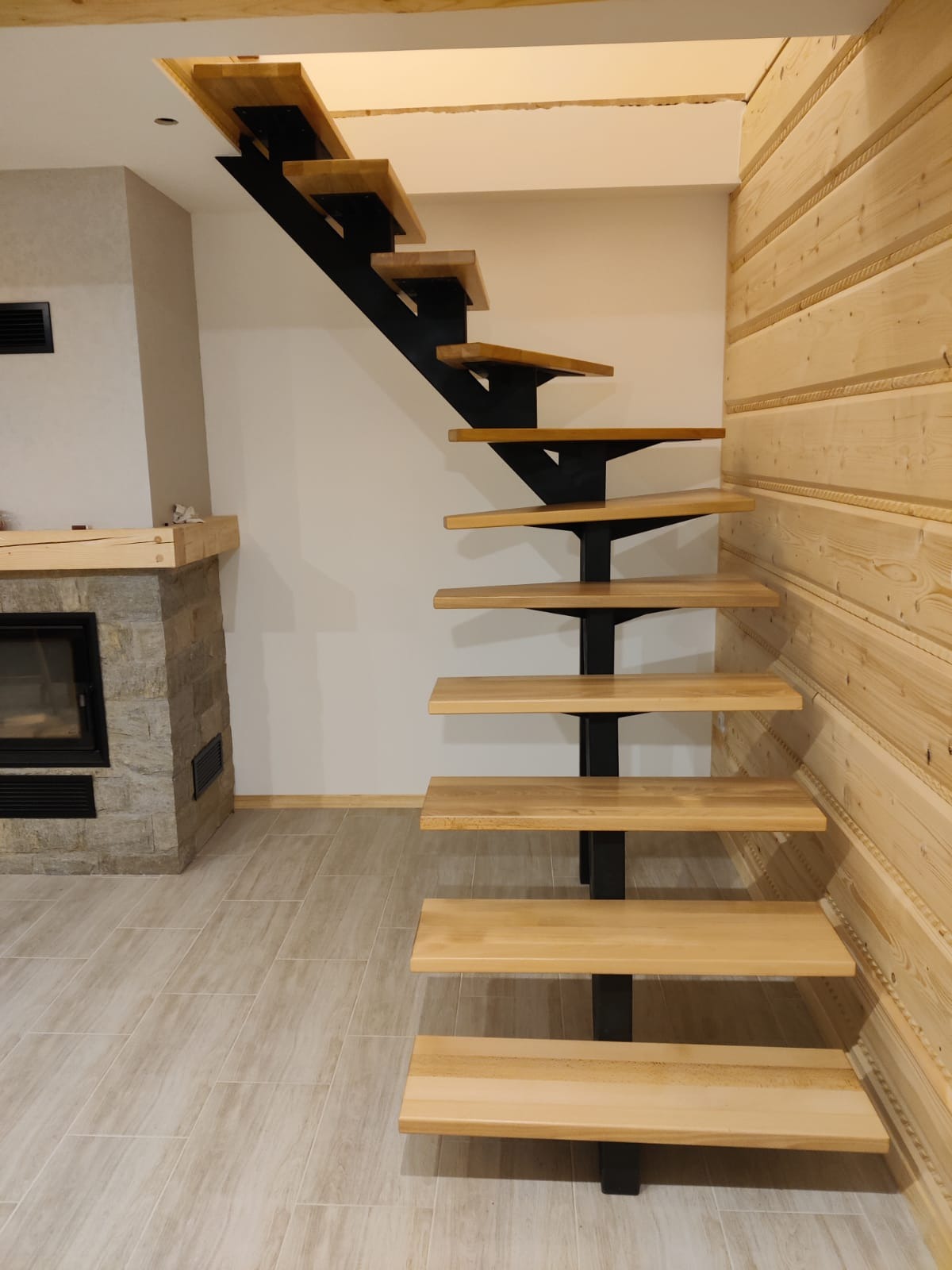 Nowoczesne, drewniane schody jednobiegowe z czarną, metalową konstrukcją, umieszczone w pomieszczeniu z drewnianą boazerią i kamiennym kominkiem.