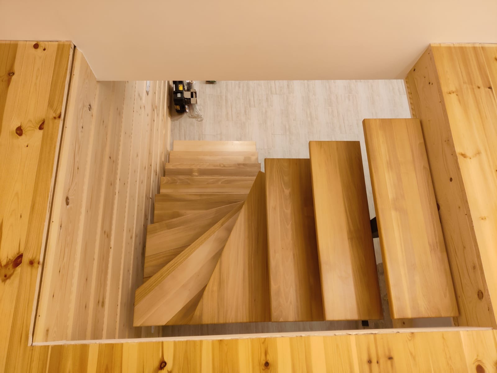 Widok z góry na drewniane schody kręte z jasnego drewna, wkomponowane w drewnianą zabudowę ścian i podłogi, z widocznym fragmentem podłogi na górze.