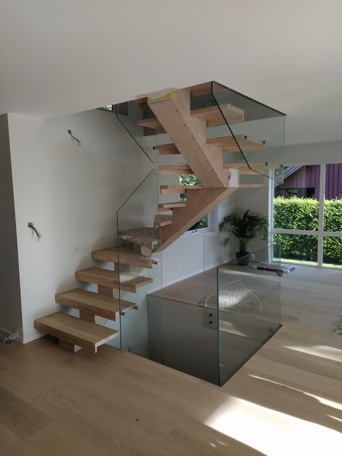 Nowoczesne schody drewniane z przezroczystą szklaną balustradą w jasnym wnętrzu domu, widoczne niedokończone detale i wystające przewody elektryczne.