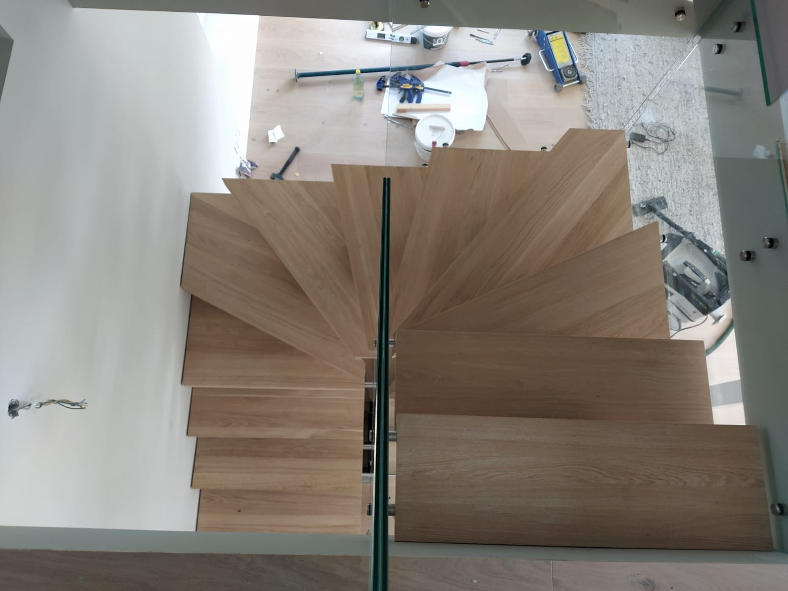 Widok z góry na spiralne drewniane schody z szklaną balustradą w trakcie montażu, narzędzia i akcesoria budowlane widoczne na podłodze w tle.