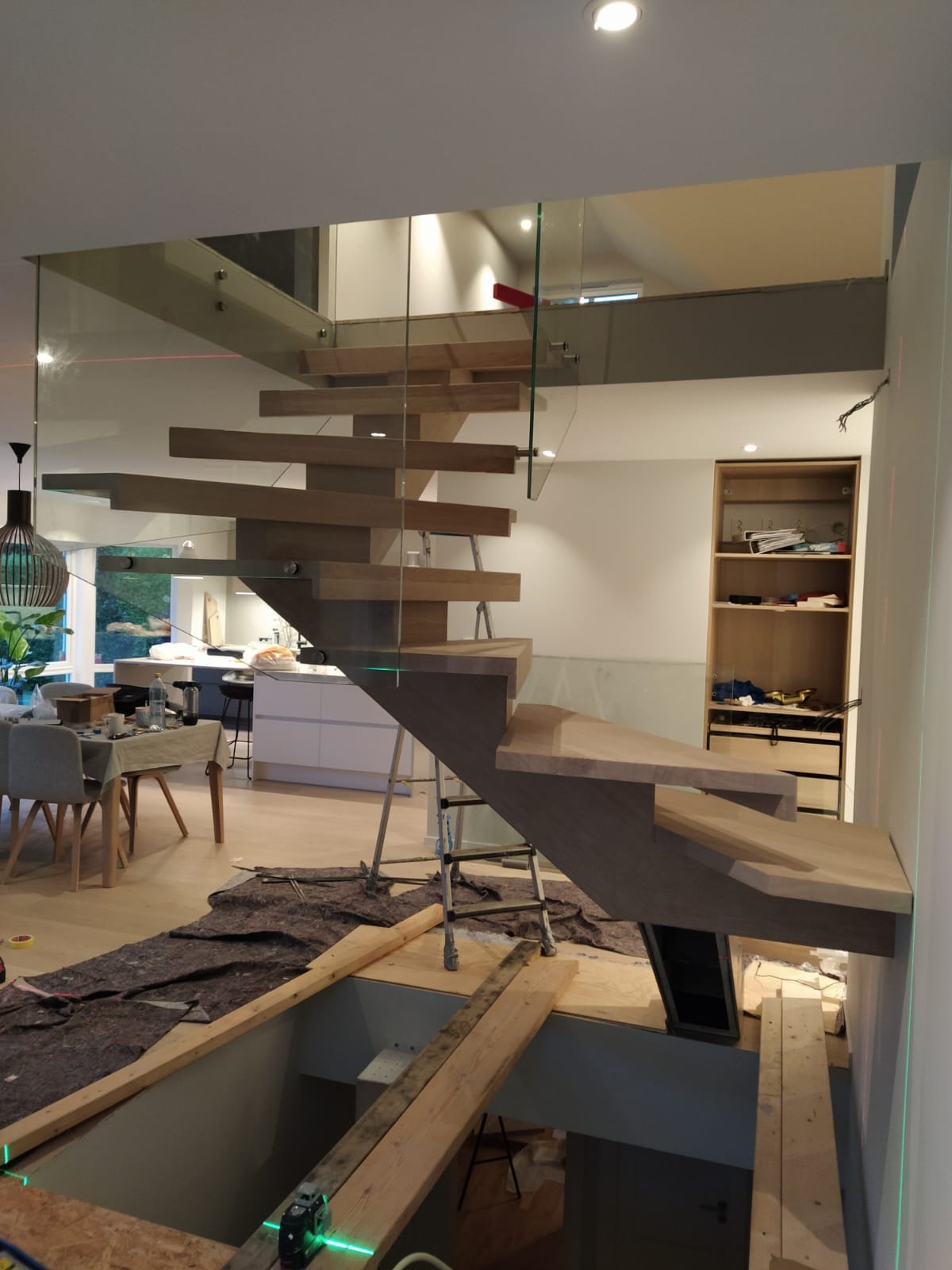 Montaż drewnianych schodów z szklaną balustradą w nowoczesnym wnętrzu, widoczne narzędzia i laserowe wyznaczniki poziomu.