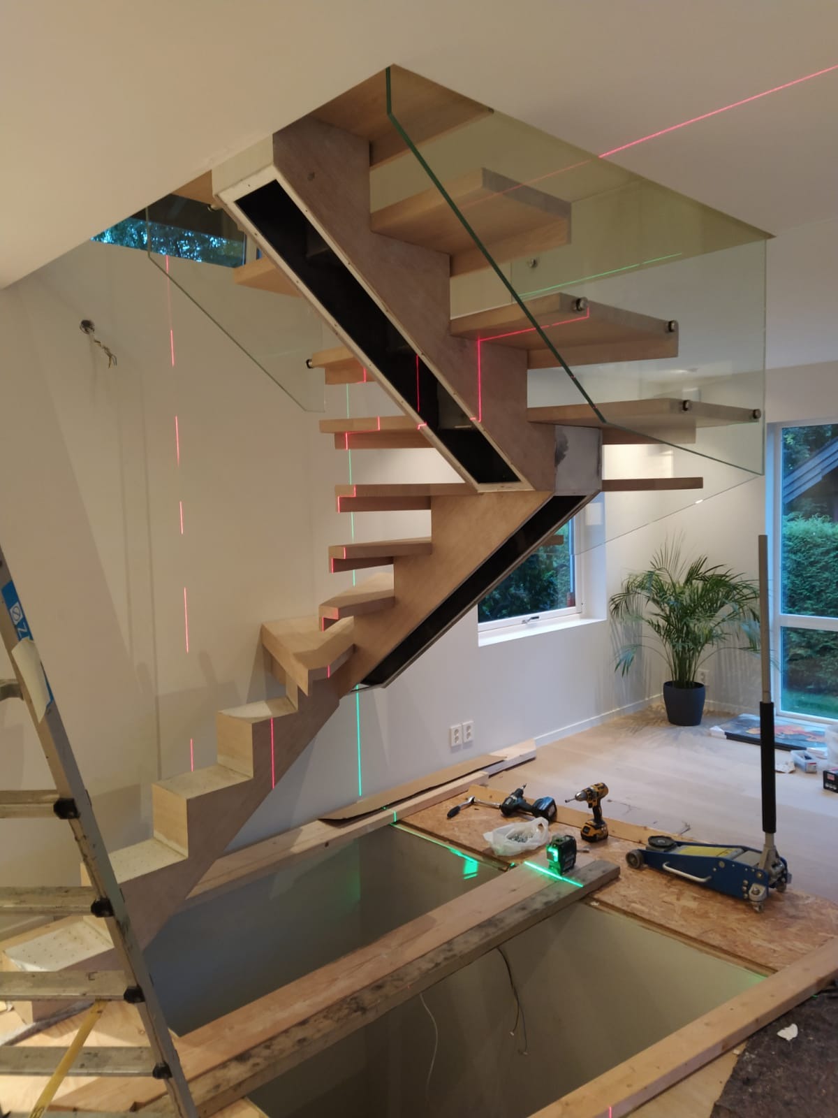 Montaż drewnianych schodów z szklaną balustradą, widoczne narzędzia i laserowe poziomice wskazujące linie cięcia, w tle okno i roślina doniczkowa.
