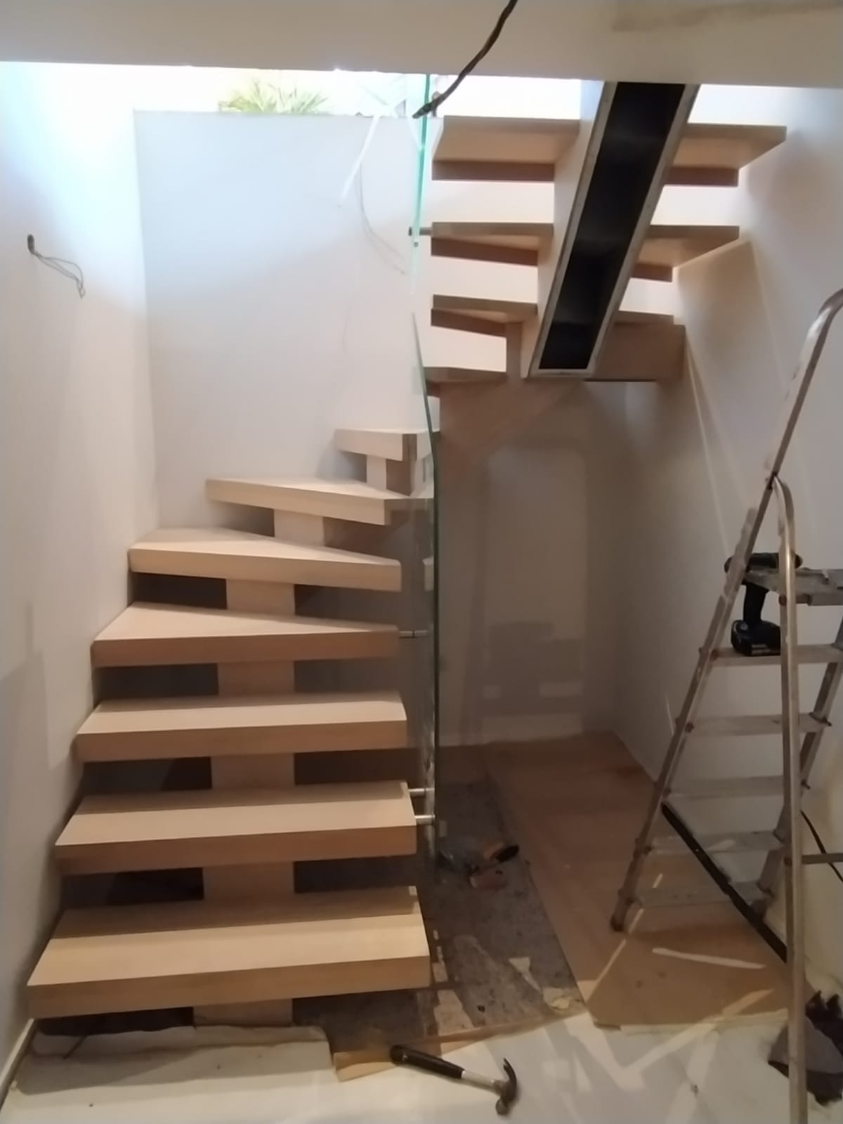 Nowoczesne schody drewniane z szklaną balustradą w trakcie montażu, widoczna drabina i narzędzia na podłodze.