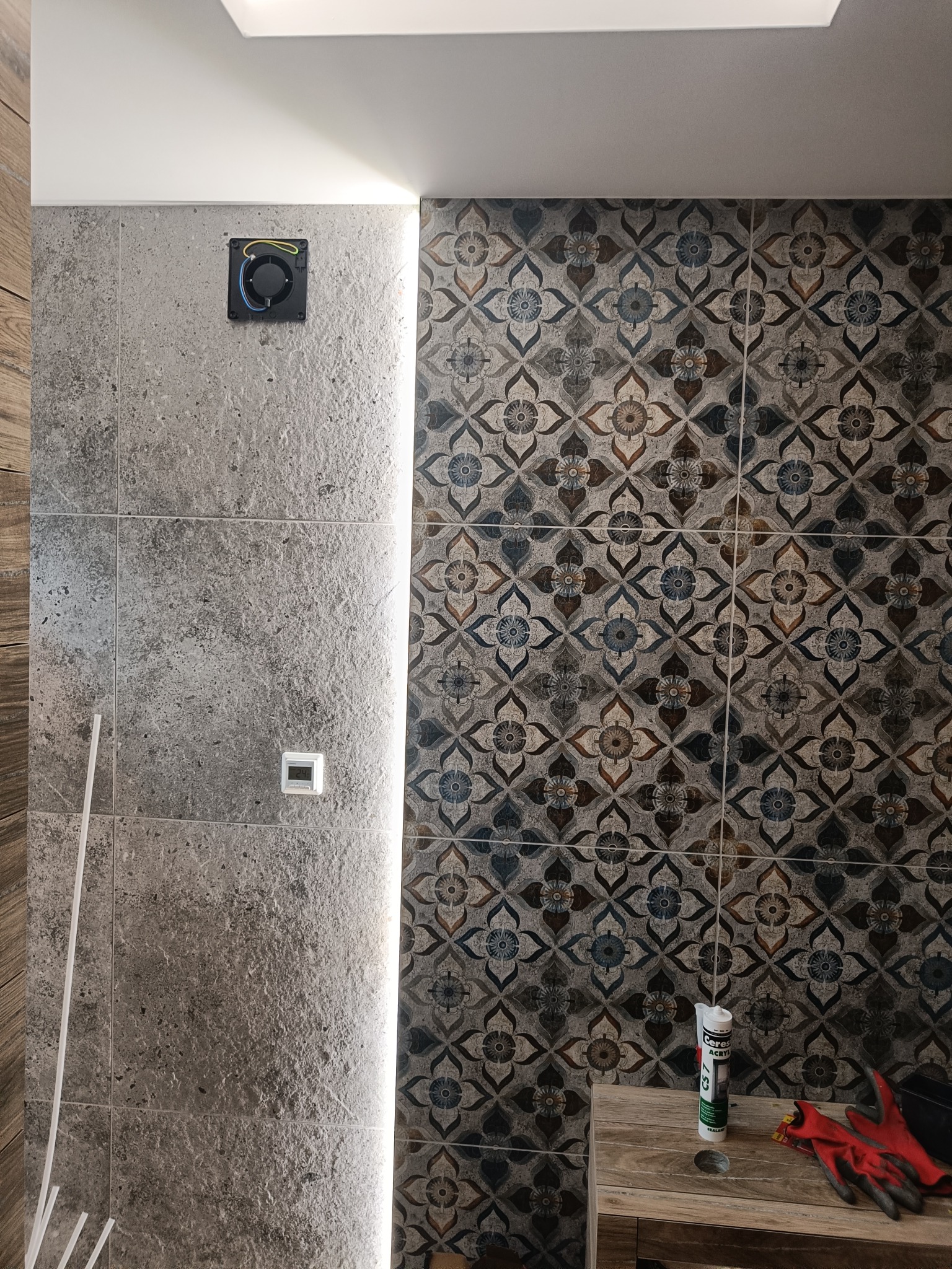 Ściana łazienki wyłożona dwoma rodzajami płytek: szare płytki imitujące kamień z czarną puszką instalacyjną i białą listwą LED oraz płytki dekoracyjne we wzory kwiatowe, obok stoi stół z tubą...