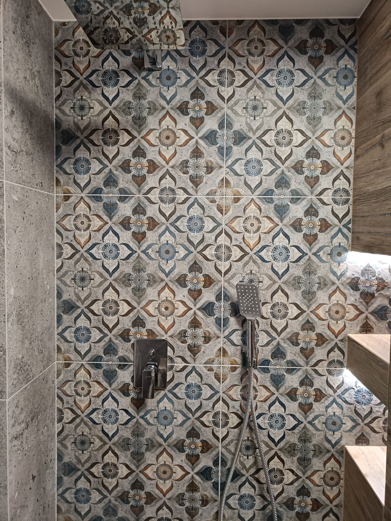 Wykończona ściana prysznicowa z ozdobnymi, wzorzystymi płytkami w odcieniach szarości, brązu i błękitu, z zamontowaną baterią i słuchawką prysznicową, widoczny fragment drewnianej obudowy.