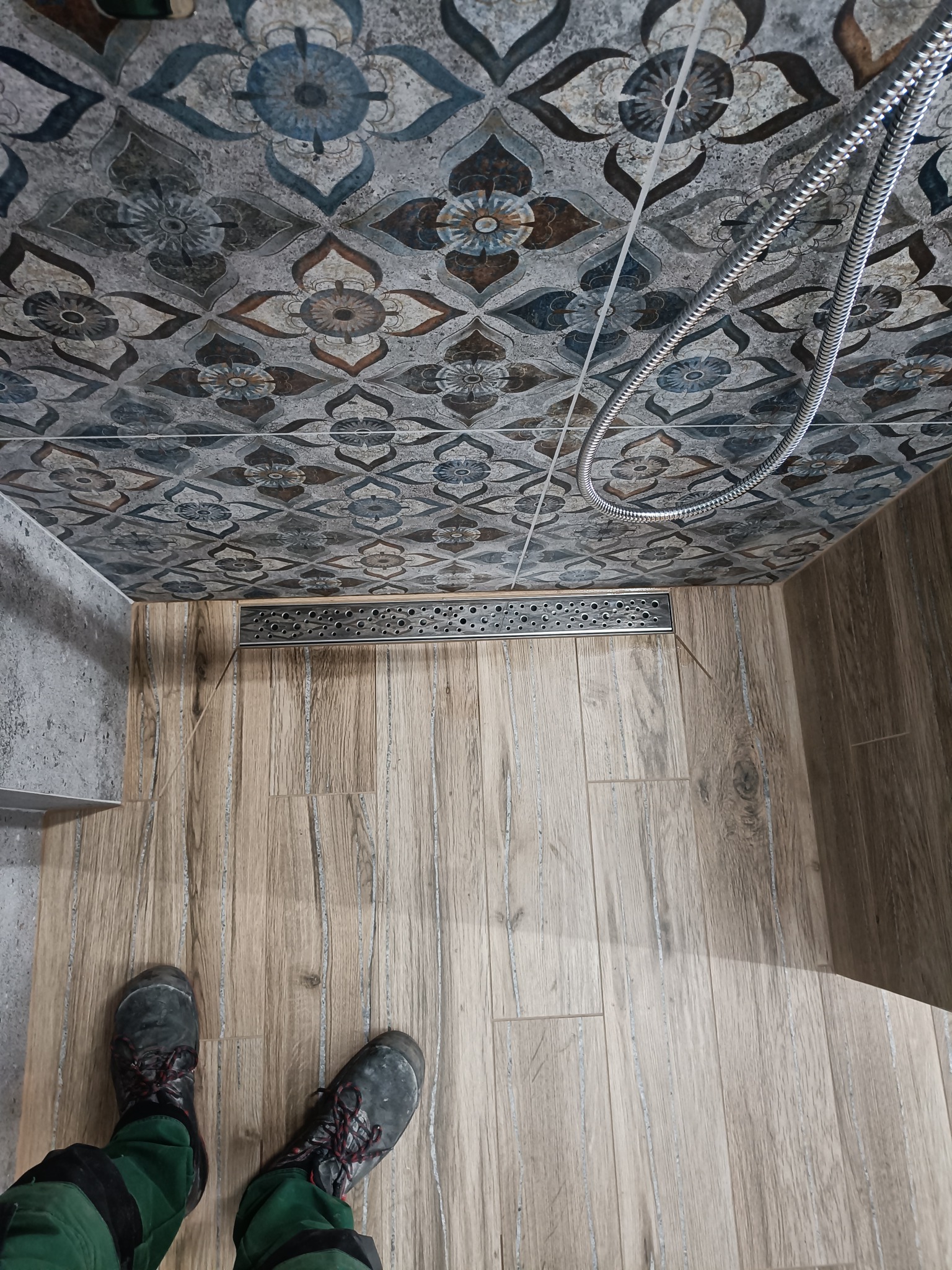 Ukończona kabina prysznicowa z odpływem liniowym, wykończona płytkami imitującymi drewno na podłodze i wzorzystymi płytkami na ścianach, widoczne buty robocze w dolnej części kadru.