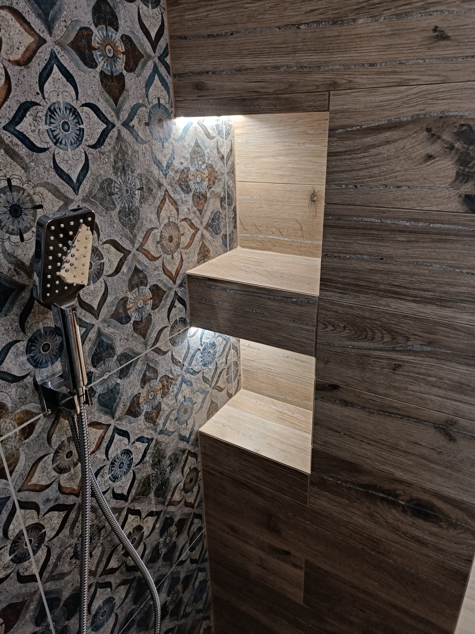 Nowoczesna łazienka z prysznicem: ściana wyłożona płytkami w stylu patchwork i imitującymi drewno, podświetlane półki w zabudowie, chromowana słuchawka prysznicowa.