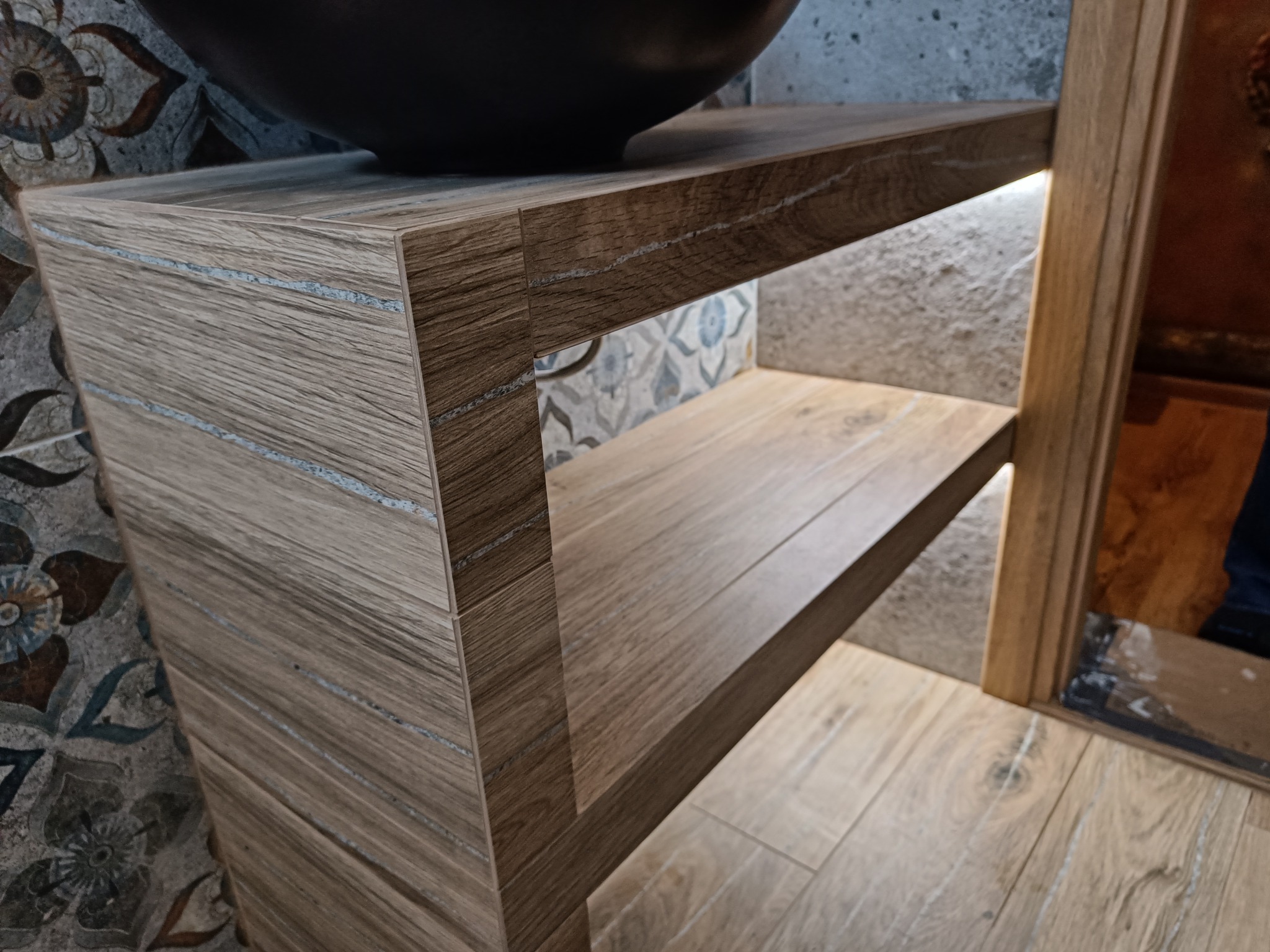 Nowoczesna umywalka nablatowa w kolorze czarnym, osadzona na drewnianym regale z oświetleniem LED, w łazience w Żabnicy z dekoracyjnymi płytkami na ścianie.