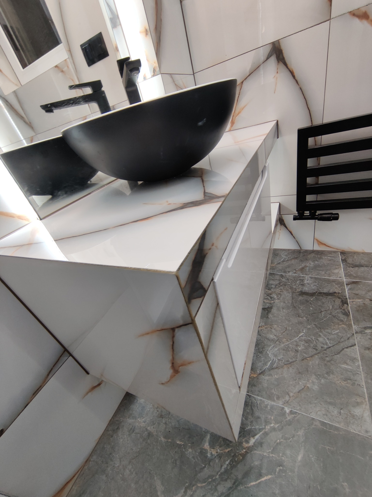 Nowoczesna łazienka z umywalką w kształcie misy, białe płytki imitujące marmur z brązowymi żyłami na ścianach i szafce pod umywalką, szare marmurowe płytki podłogowe oraz czarny grzejnik drabinkowy.