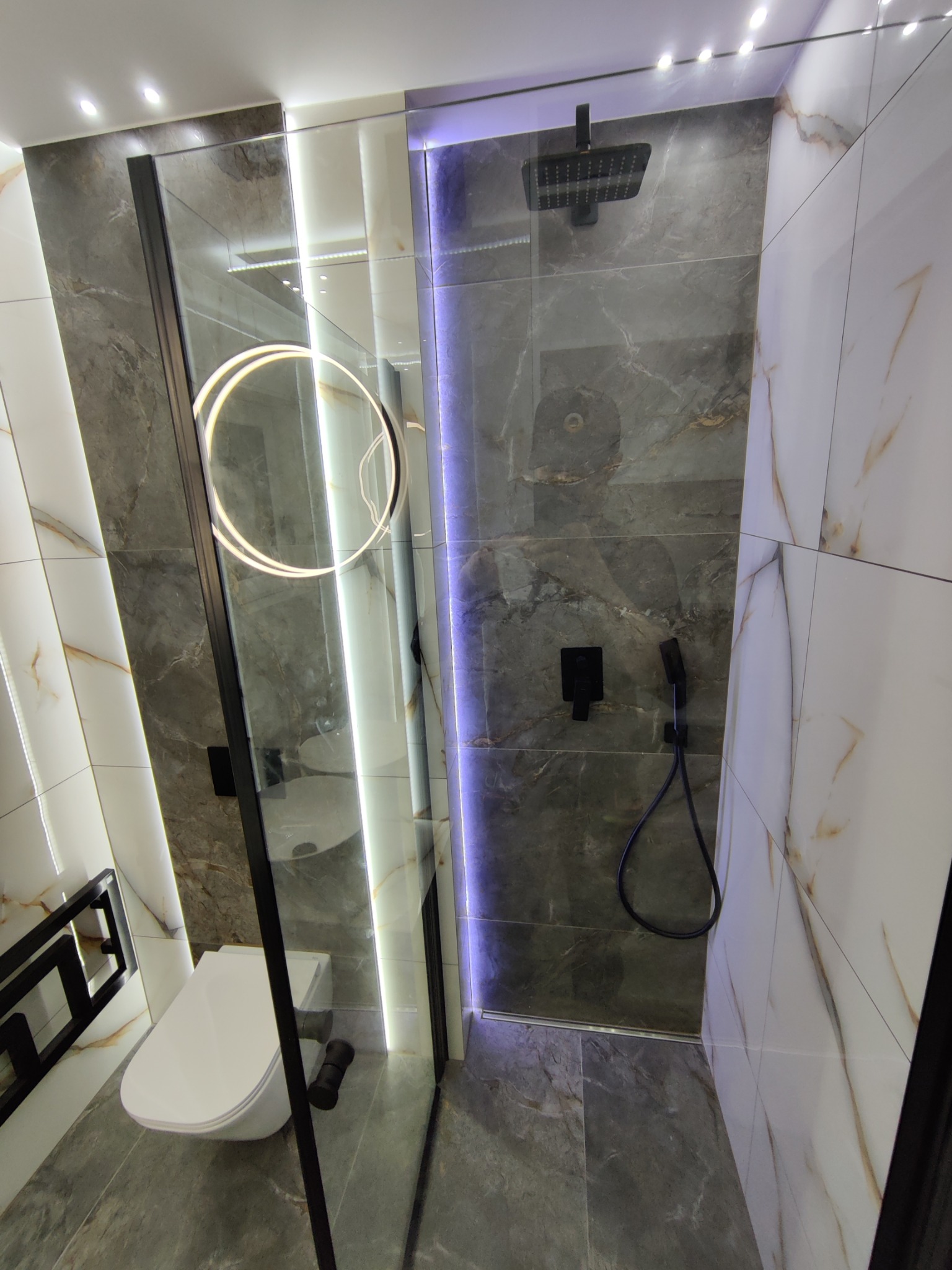 Nowoczesna łazienka z kabiną prysznicową typu walk-in, wykończona płytkami imitującymi marmur w odcieniach szarości i bieli, z czarną armaturą i oświetleniem LED.