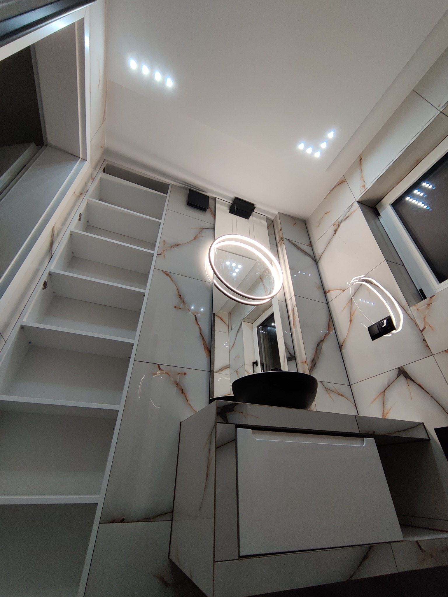 Nowoczesna łazienka z marmurowymi płytkami, okrągłym lustrem z podświetleniem LED, czarną umywalką nablatową i wysoką białą szafką z otwartymi półkami po lewej stronie.