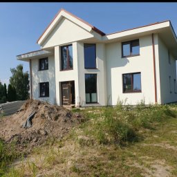 Viktermo - Nowoczesny, dwukondygnacyjny dom z jasną elewacją i brązowymi ramami okiennymi. Na pierwszym planie hałda ziemi i materiały budowlane, sugerujące trwające prace wykończeniowe.