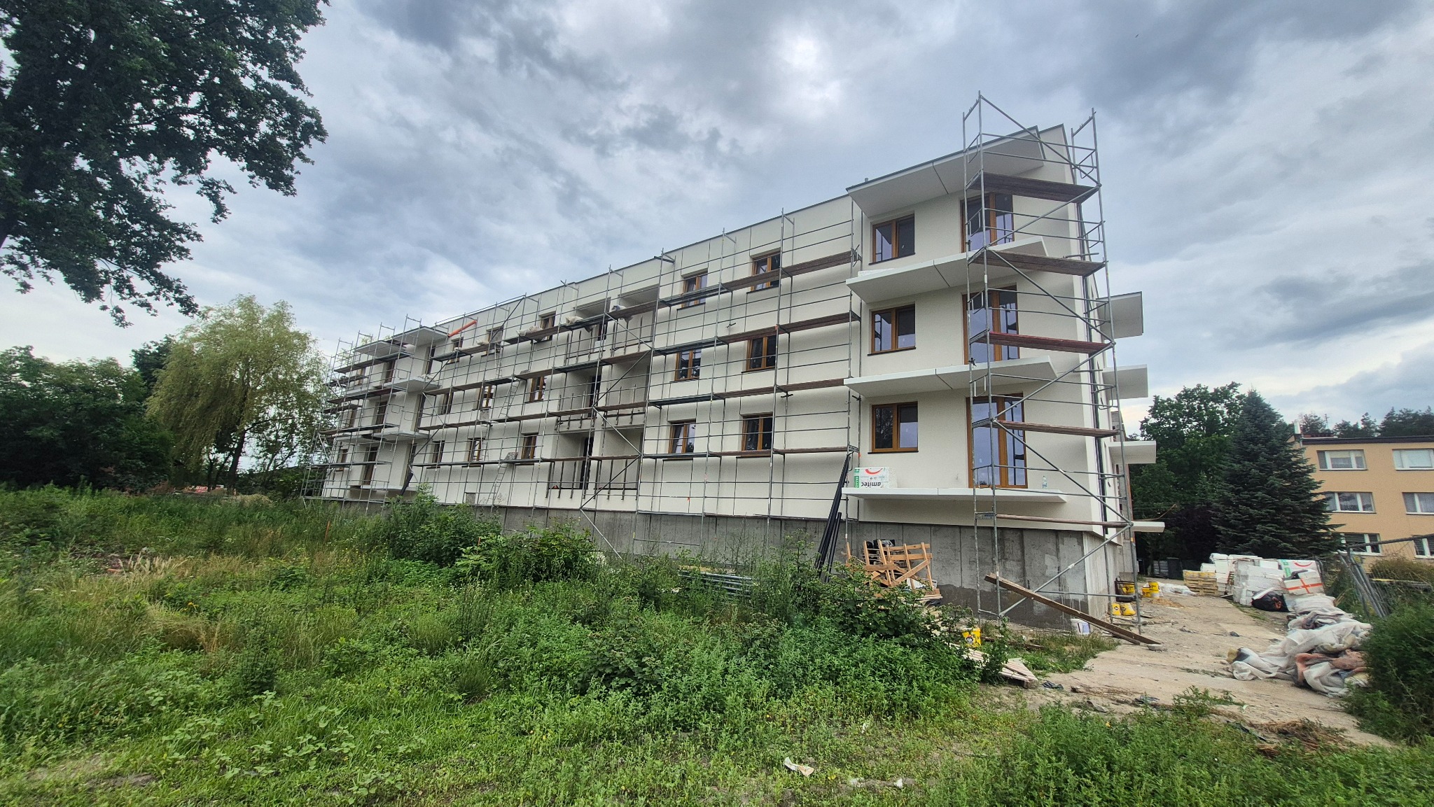 Wielopiętrowy budynek w trakcie ocieplania elewacji, z widocznymi rusztowaniami i częściowo otynkowaną fasadą. Widoczne materiały budowlane i otoczenie zieleni.