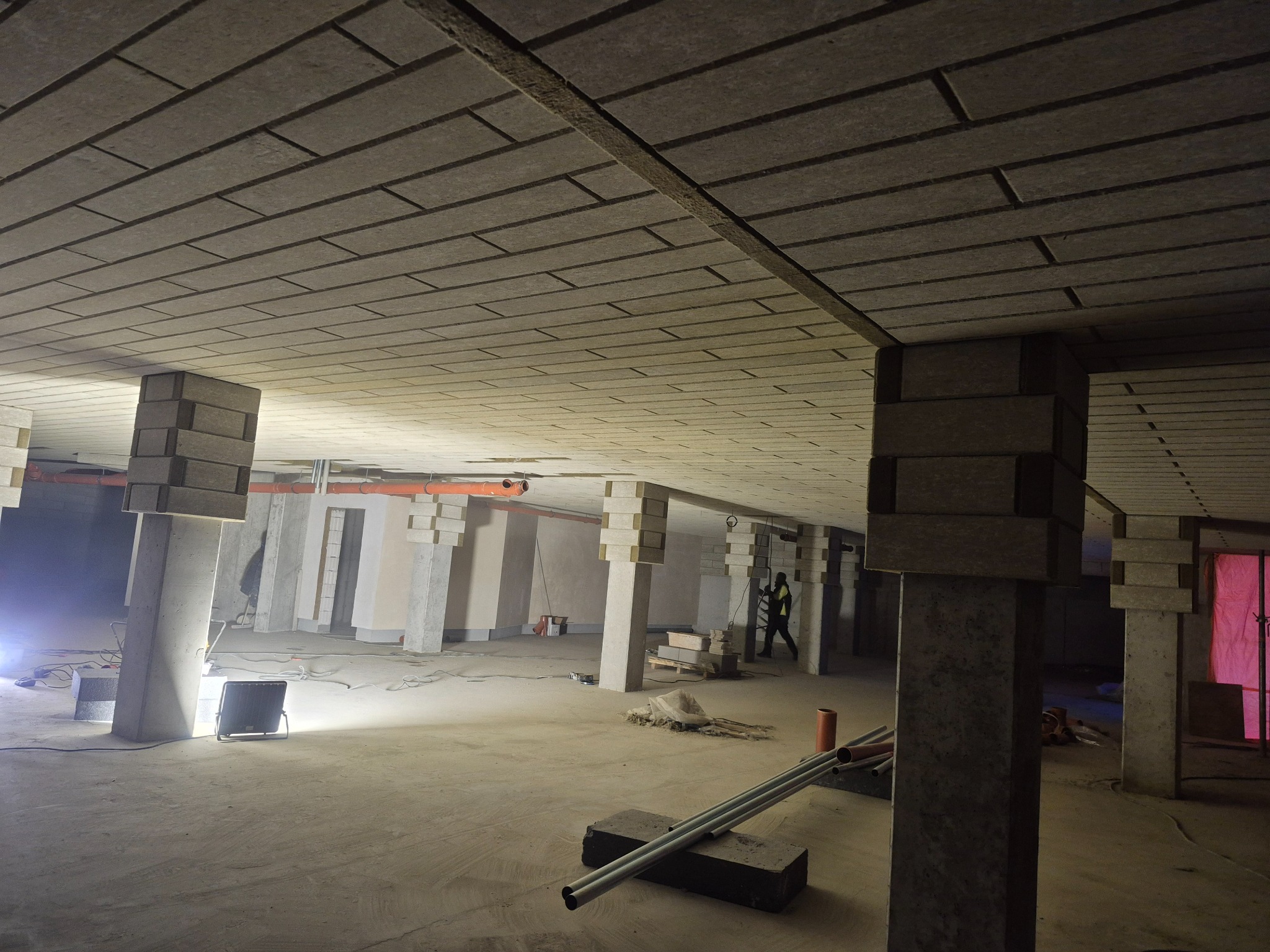 Wykończone wnętrze hali z izolowanym stropem kasetonowym i kolumnami. Widoczne elementy instalacji i oświetlenie budowlane. Prace wykończeniowe w toku.