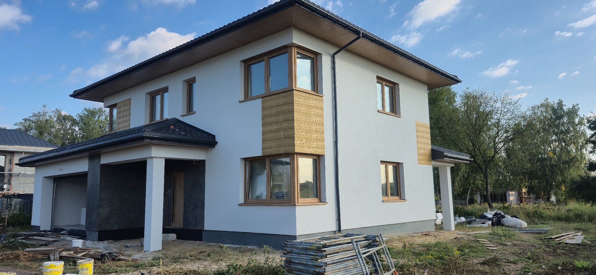 Dwukondygnacyjny dom z białą elewacją i drewnianymi akcentami. Widoczne rusztowania i materiały budowlane sugerują prace wykończeniowe.