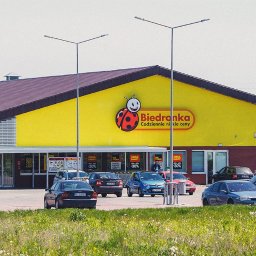 KGDI ZARZĄDZANIE SPÓŁKA Z OGRANICZONĄ ODPOWIEDZIALNOŚCIĄ - Żółty budynek Biedronki z parkującymi samochodami w słoneczny dzień. Widoczny szyld sklepu i zaparkowane auta. Trawa i drzewa w tle.