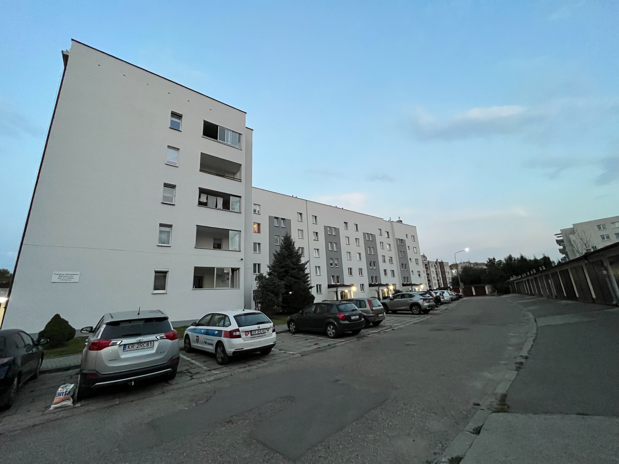 Nowoczesny budynek mieszkalny z białymi ścianami, balkonami i szarymi akcentami, otoczony parkingiem z kilkoma samochodami, sfotografowany o zmierzchu.