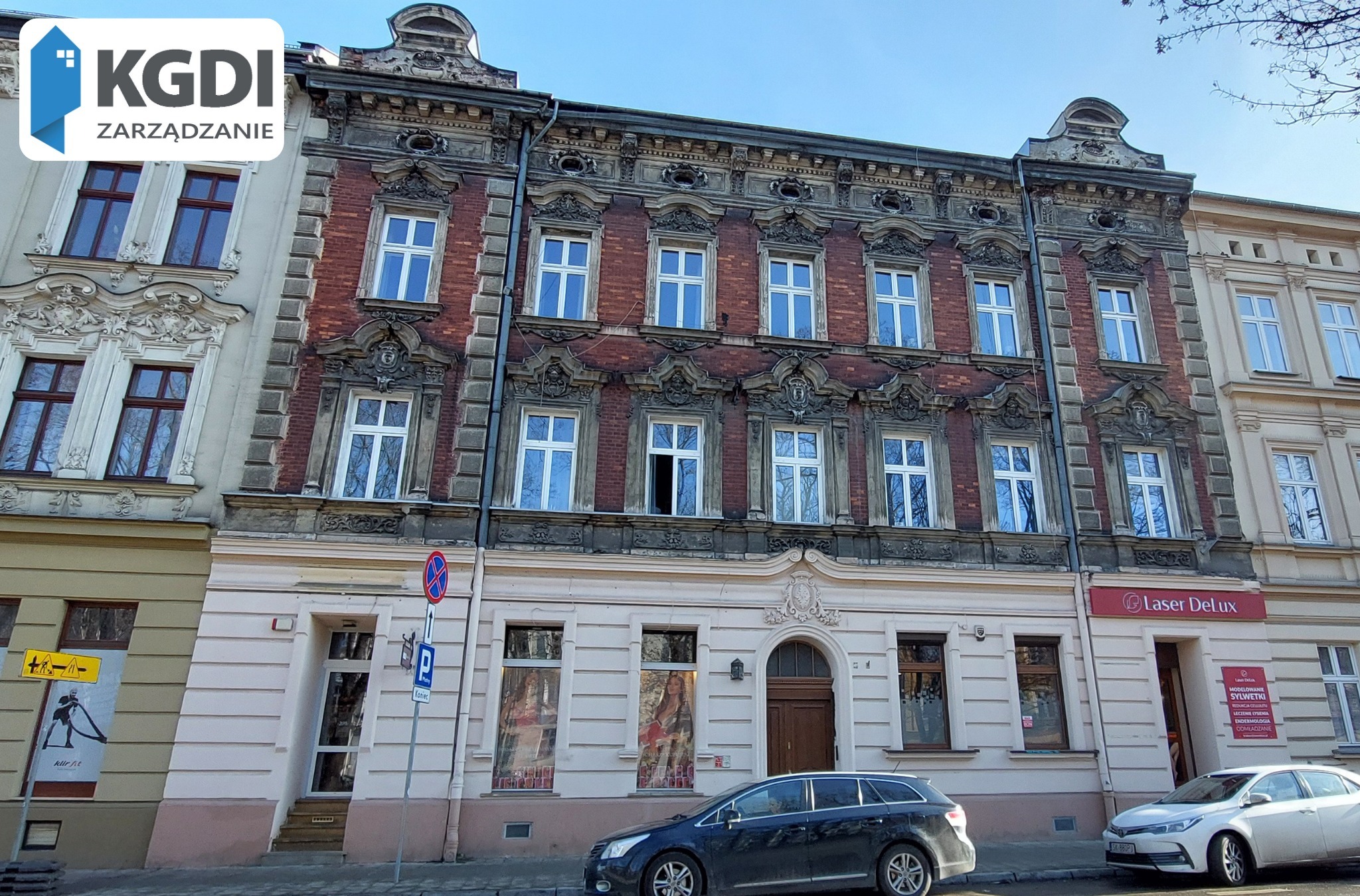Fasada kamienicy z elementami secesyjnymi, z widocznym logo firmy KGDI Zarządzanie w lewym górnym rogu, zaparkowane samochody na pierwszym planie, oraz szyld Laser DeLux po prawej stronie.