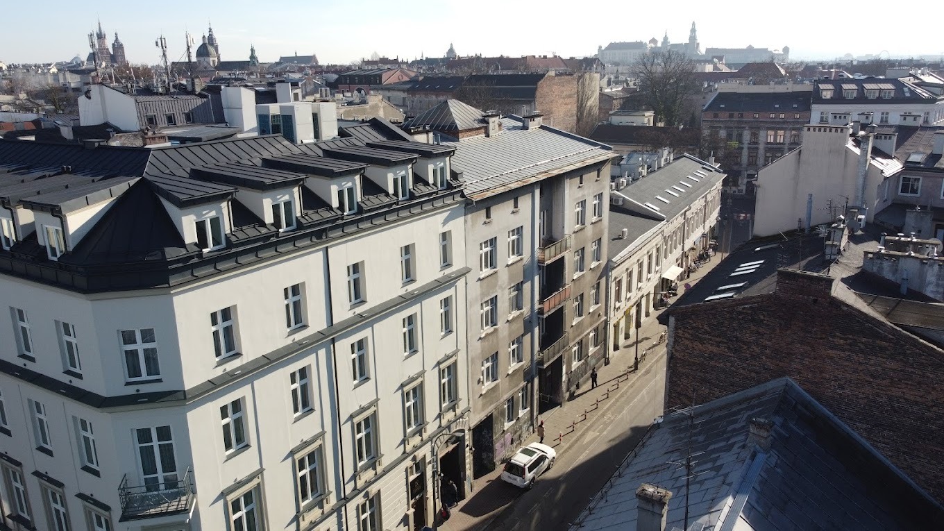 Panorama miejska z widokiem na odnowione kamienice z ciemnymi dachami i jasnymi elewacjami w słoneczny dzień, w tle historyczne budowle Krakowa.