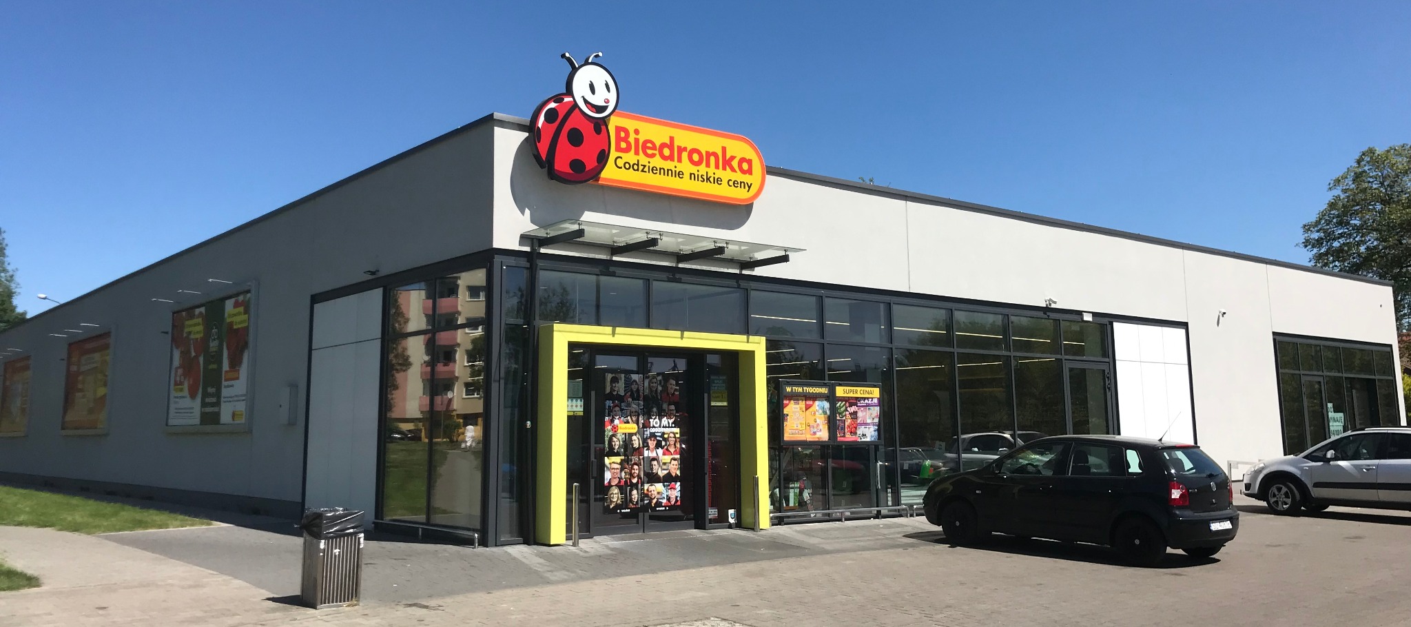 Fasada sklepu Biedronka w słoneczny dzień, z widocznym logo i wejściem z żółtą ramą. Na parkingu zaparkowane samochody, a na ścianie plakaty reklamowe.