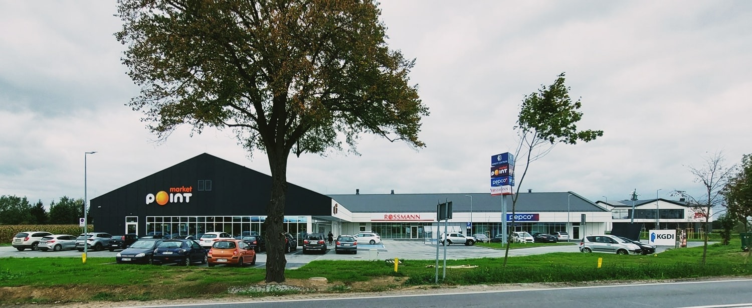 Szeroki kadr na budynek centrum handlowego Market Point z parkingiem i zielenią, widziany z perspektywy ulicy w pochmurny dzień. Widoczne logo Rossmann i Pepco.