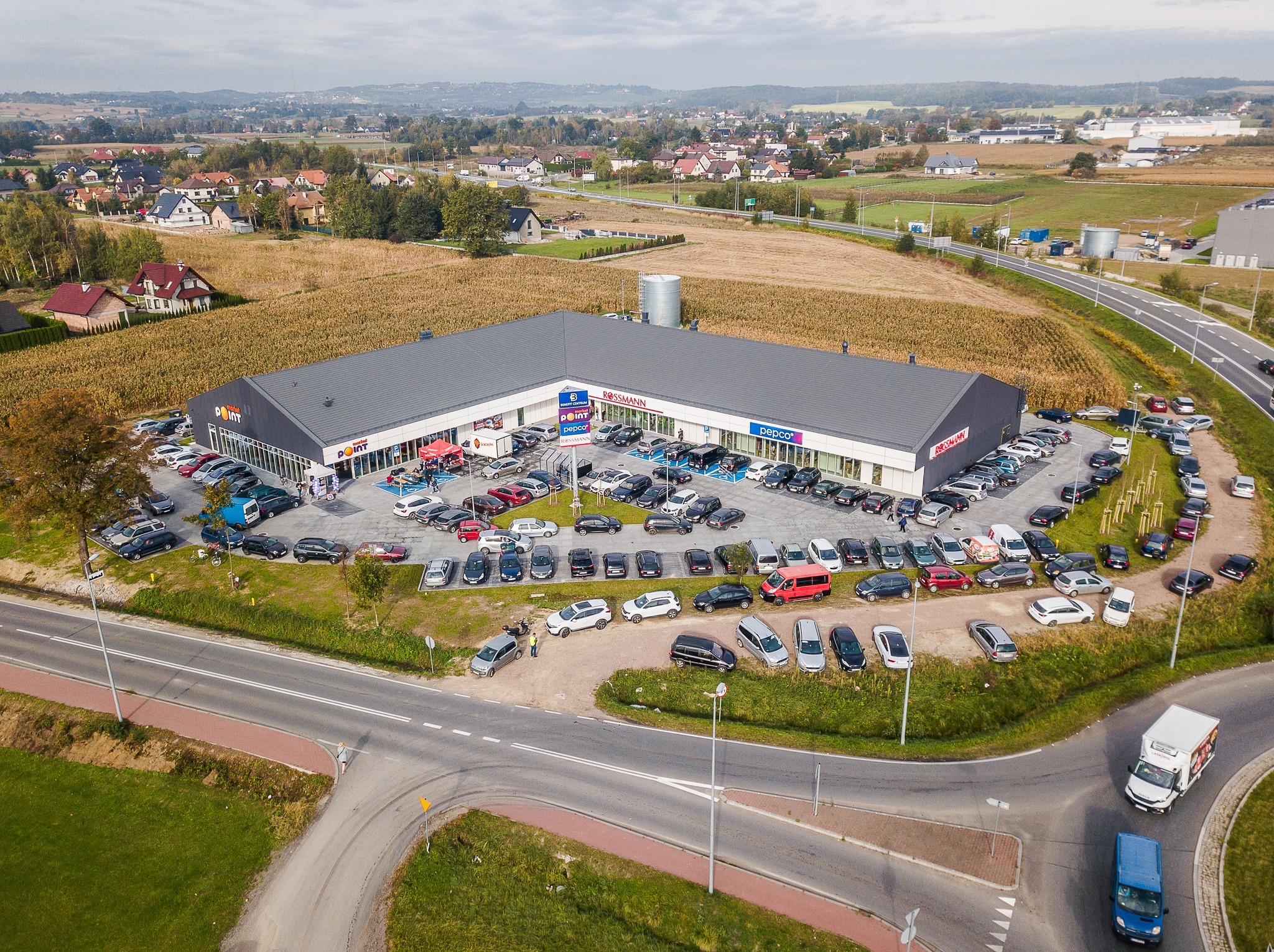 Zdjęcie z lotu ptaka przedstawia budynek centrum handlowego z parkingiem pełnym samochodów, otoczonego polami i zabudową mieszkalną. Widoczny ruch na drogach.