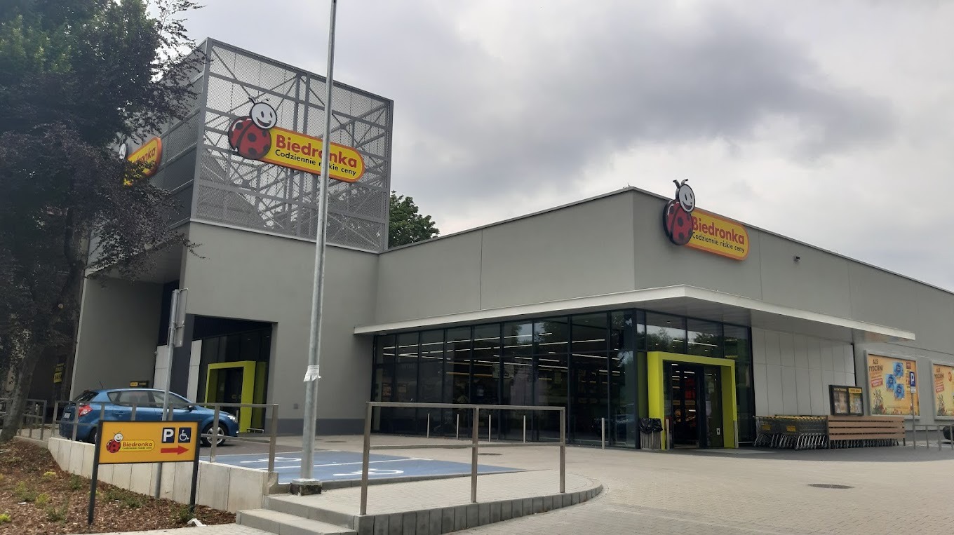 Zewnętrzny widok nowoczesnego budynku sklepu Biedronka z charakterystycznym logo biedronki, parkingiem i zielenią w tle, uchwycony w pochmurny dzień.