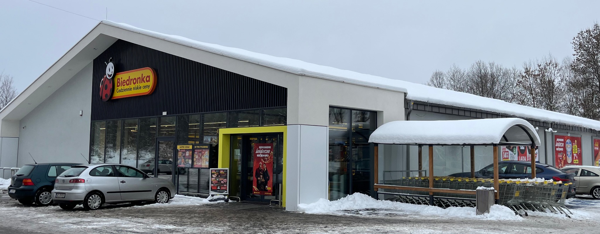 Zimowy krajobraz z budynkiem sklepu Biedronka, pokrytym śniegiem. Widoczne zaparkowane samochody i wózki sklepowe pod zadaszeniem.