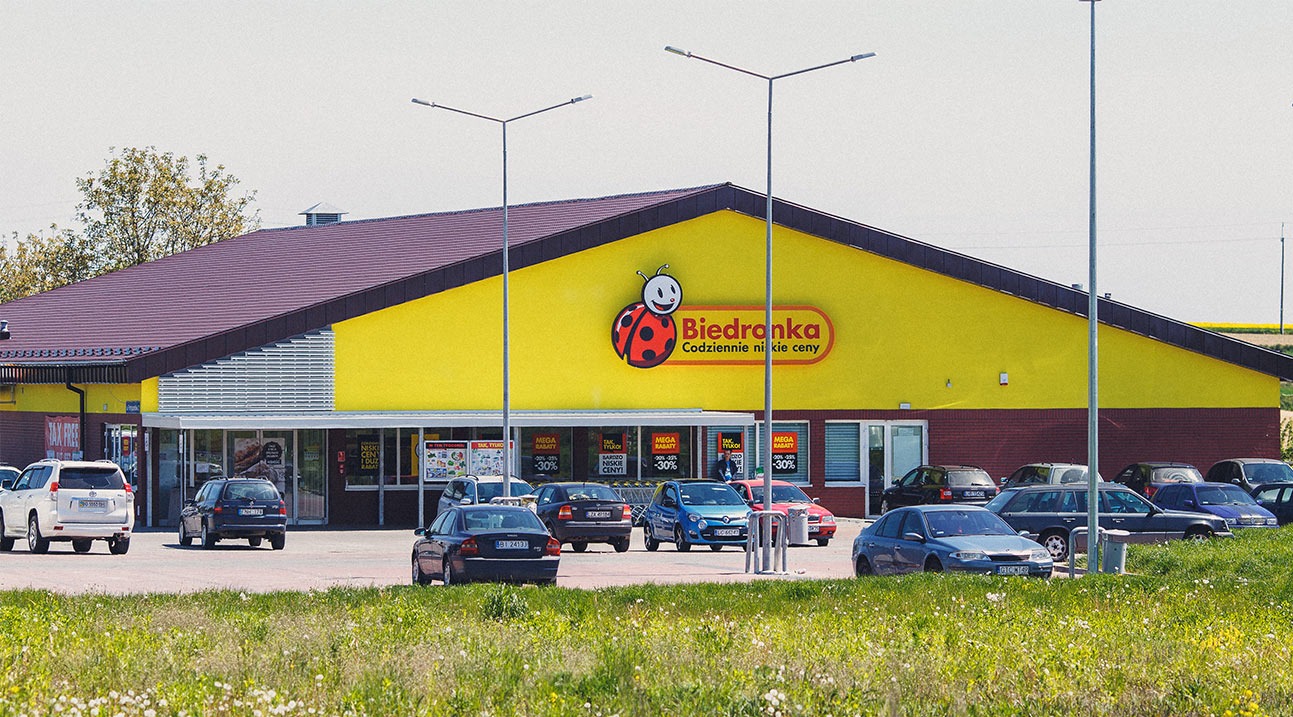 Żółty budynek Biedronki z parkującymi samochodami w słoneczny dzień. Widoczny szyld sklepu i zaparkowane auta. Trawa i drzewa w tle.