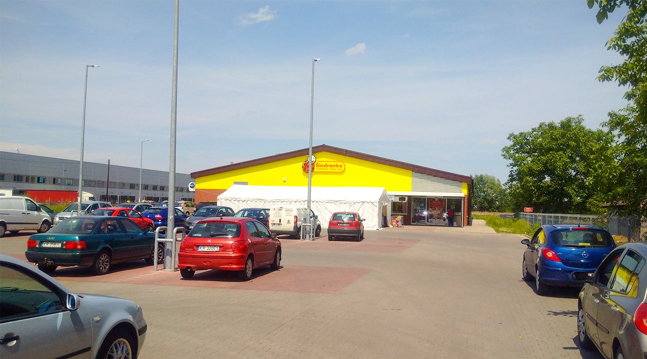 Widok na parking przed budynkiem sklepu Biedronka z żółtą fasadą i białym namiotem, w tle budynek magazynowy i błękitne niebo.