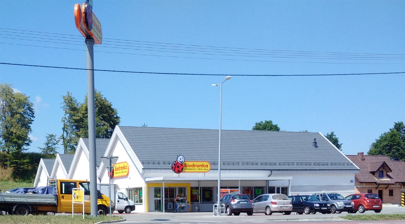 Budynek sklepu Biedronka z szarym dachem i żółtym logo na tle błękitnego nieba. Na parkingu zaparkowane samochody, a przed budynkiem wózki sklepowe.