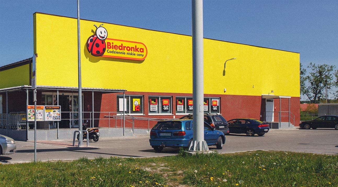 Żółto-czerwony budynek Biedronki z parkującymi samochodami i zielonym trawnikiem. Widoczne plakaty promocyjne w oknach i charakterystyczne logo sieci.