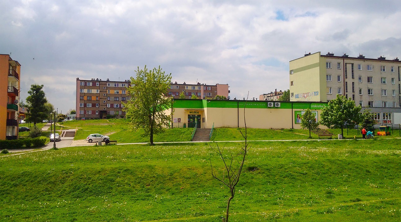 Osiedle mieszkaniowe z budynkami wielorodzinnymi, zielonym terenem i sklepem Stokrotka. Widok na budynki z trawnika, pochmurne niebo.
