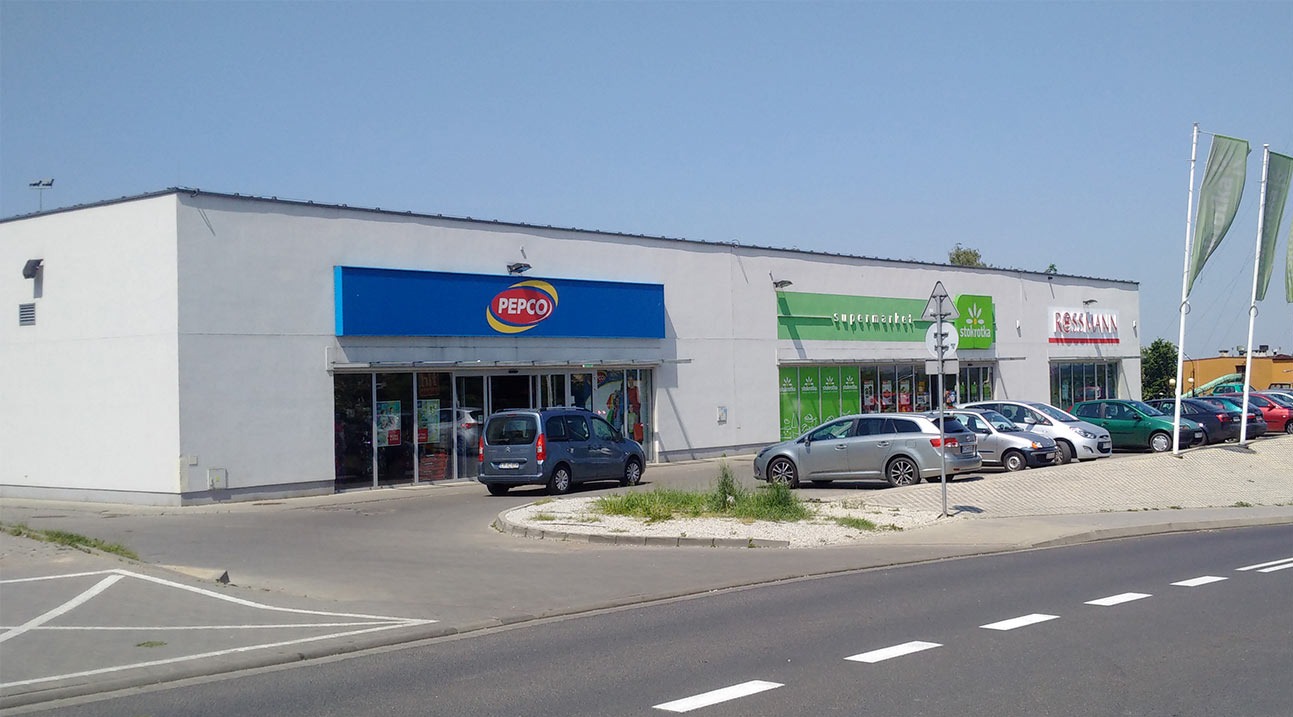 Budynek handlowy z kilkoma sklepami: Pepco, Stokrotka, Rossmann. Na parkingu zaparkowane samochody. Widok z ulicy w słoneczny dzień.