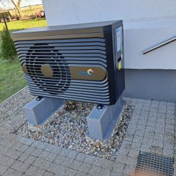 KG INSTAL Sp. z o. o. - Czarna jednostka zewnętrzna z logo FOX, zamontowana na betonowych podstawach na tle jasnej ściany budynku, widoczne kamyki i kostka brukowa.