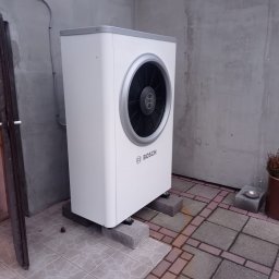 KG INSTAL Sp. z o. o. - Biała pompa ciepła Bosch zamontowana na betonowych bloczkach przy szarej ścianie budynku, widoczne przewody i fragment drzwi.