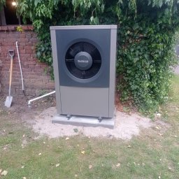 KG INSTAL Sp. z o. o. - Zewnętrzna jednostka pompy ciepła marki Buderus, zainstalowana na betonowej podstawie przy ceglanym budynku z pnącą roślinnością, obok stoi łopata.