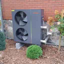 Zewnętrzna jednostka dwuwentylatorowa w kolorze grafitowym, zamontowana na betonowych podstawach przy ceglanym budynku z elementami kamiennymi, otoczona korą i niskimi krzewami.