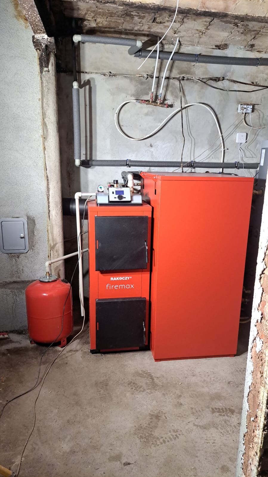 Pomieszczenie kotłowni z zamontowanym piecem Rakoczy firemax i czerwonym zbiornikiem wyrównawczym, widoczne rury i instalacja na ścianie.