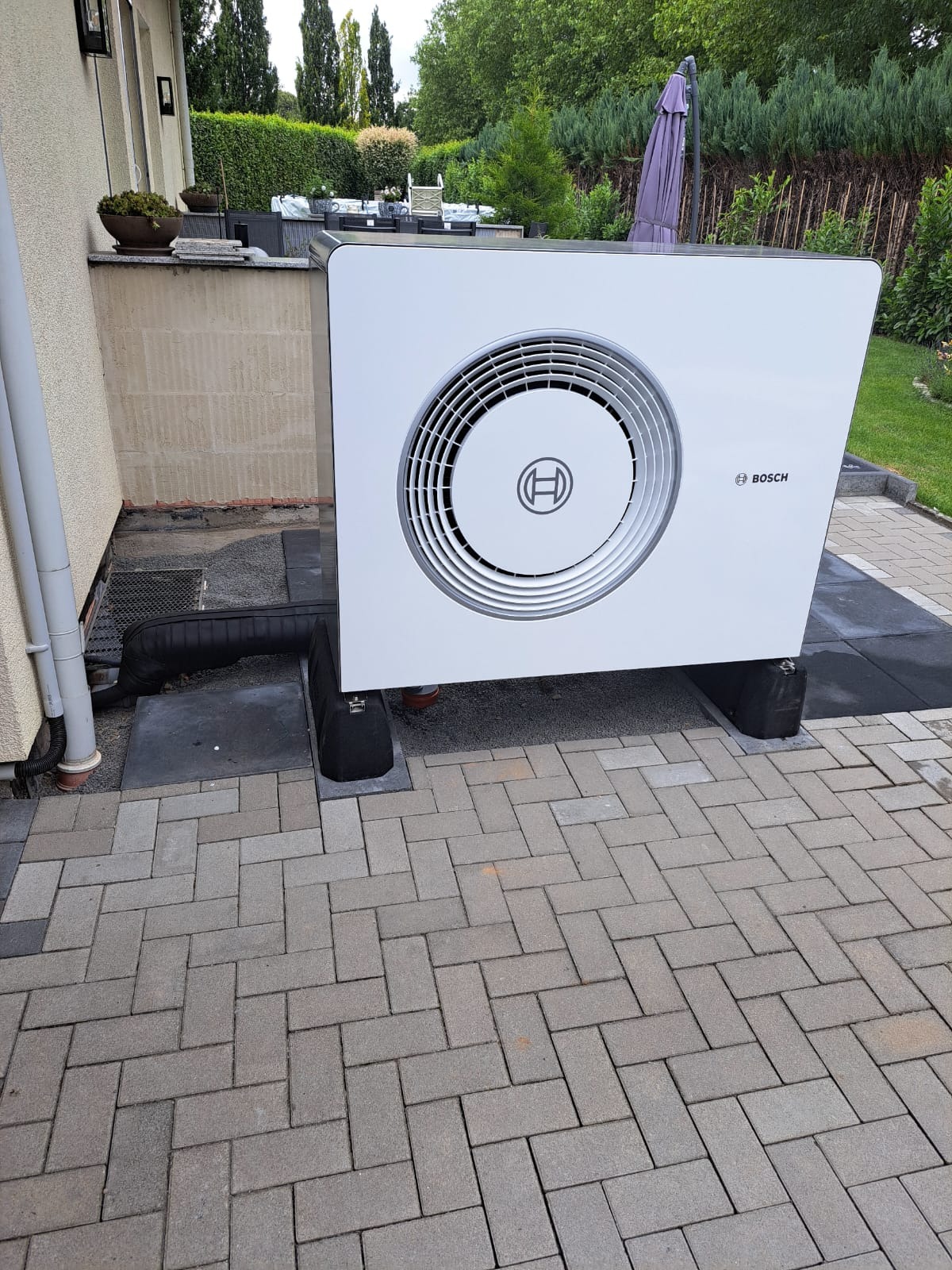 Biała jednostka zewnętrzna z logo Bosch, zainstalowana na kostce brukowej obok budynku z widocznymi elementami instalacji odprowadzającej wodę.