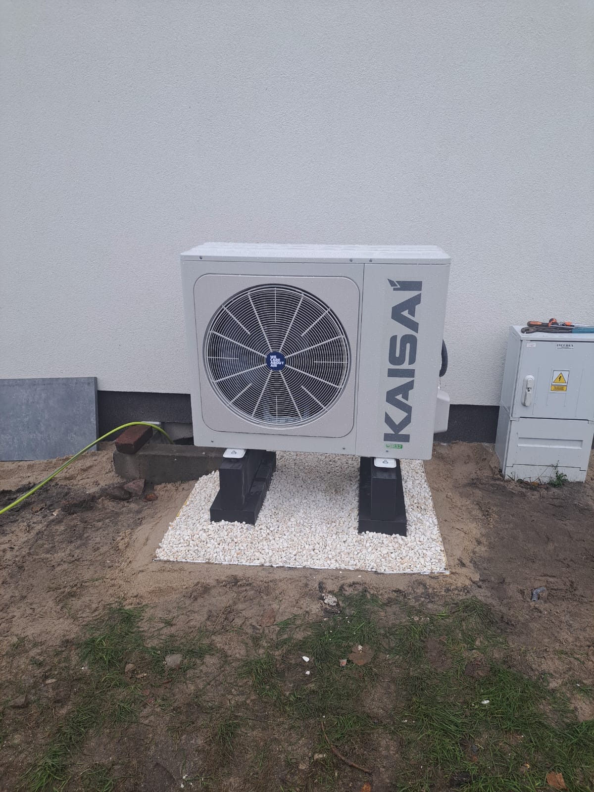 Zewnętrzna jednostka klimatyzacyjna Kaisai ustawiona na czarnych podstawkach na białym żwirze, obok skrzynka elektryczna, widoczny fragment zielonego węża.