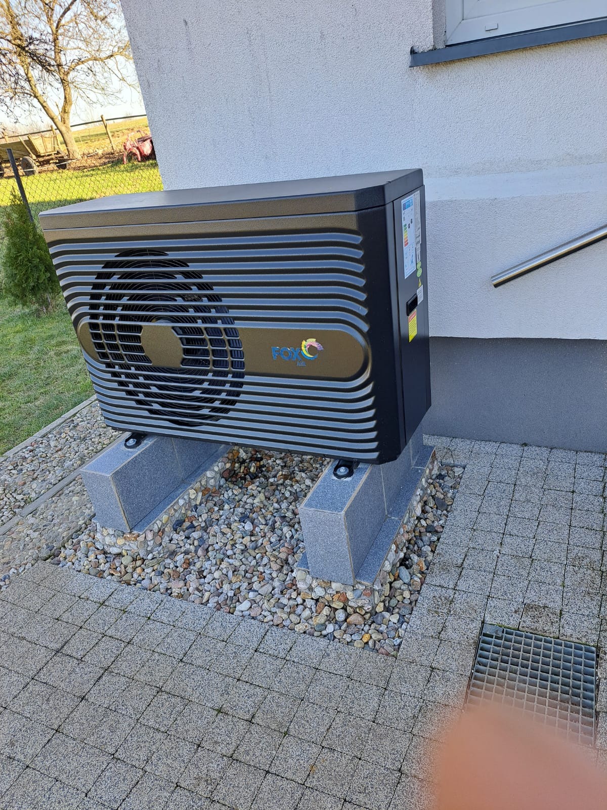 Czarna jednostka zewnętrzna z logo FOX, zamontowana na betonowych podstawach na tle jasnej ściany budynku, widoczne kamyki i kostka brukowa.