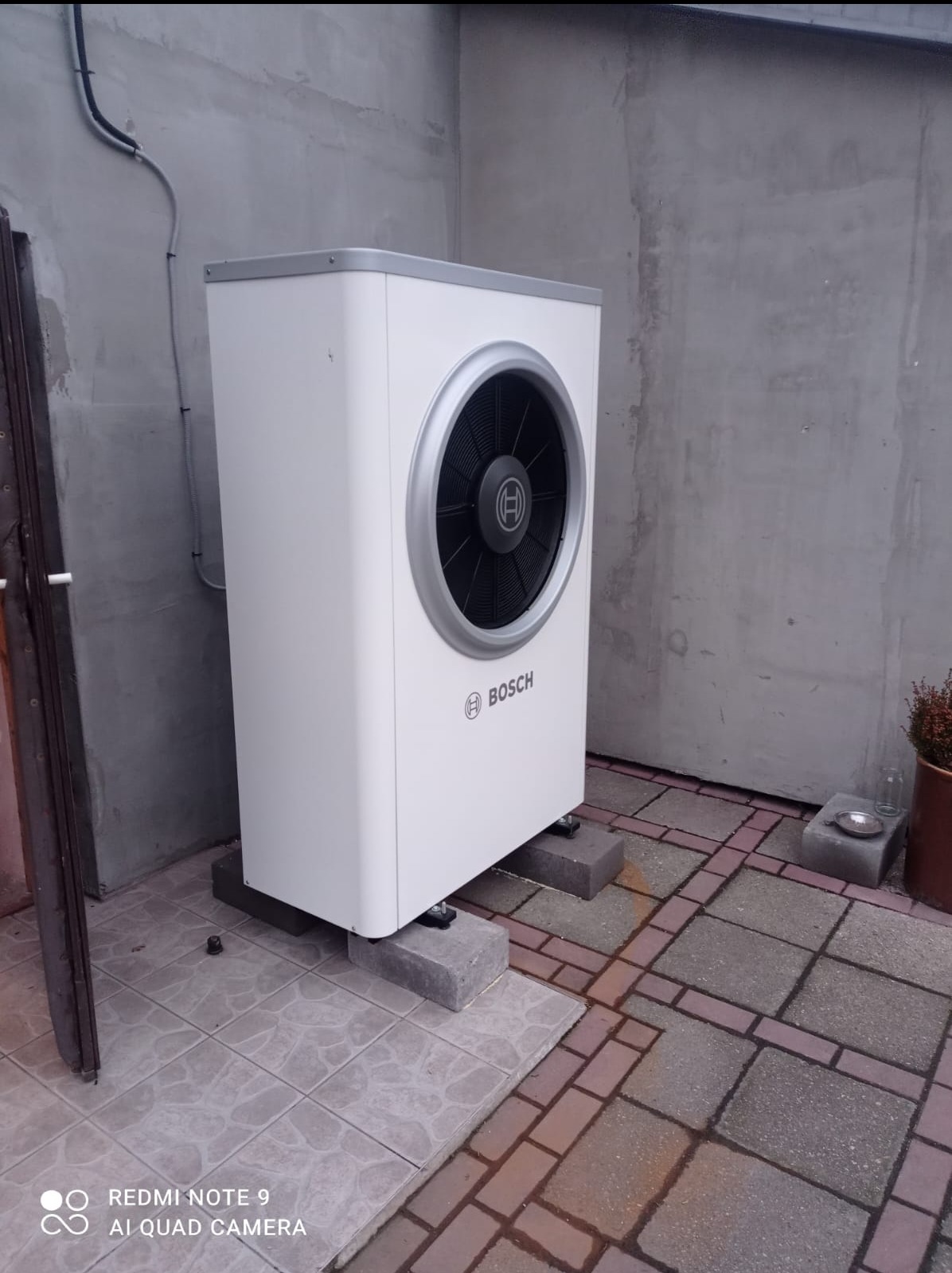 Biała pompa ciepła Bosch zamontowana na betonowych bloczkach przy szarej ścianie budynku, widoczne przewody i fragment drzwi.
