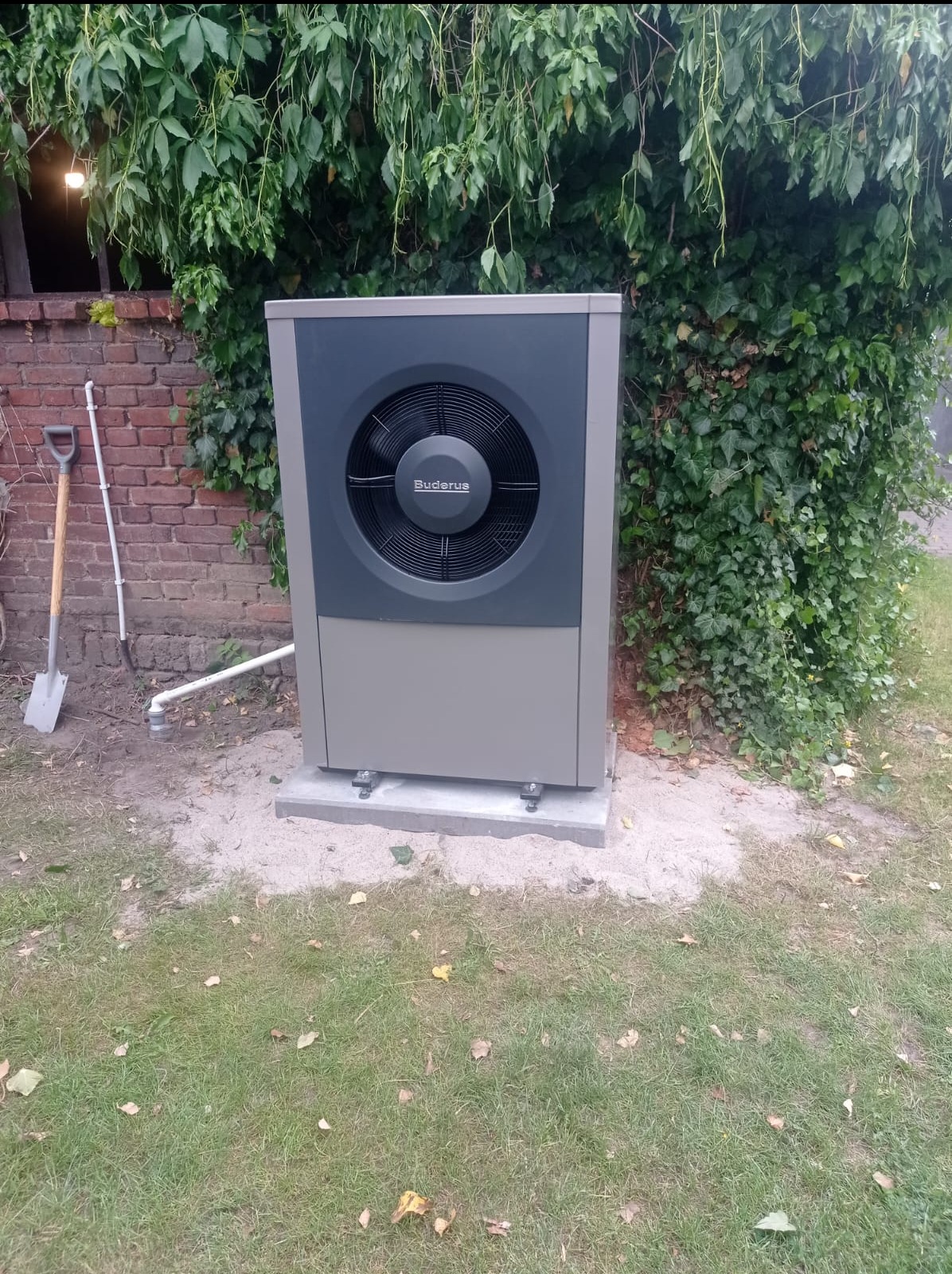 Zewnętrzna jednostka pompy ciepła marki Buderus, zainstalowana na betonowej podstawie przy ceglanym budynku z pnącą roślinnością, obok stoi łopata.