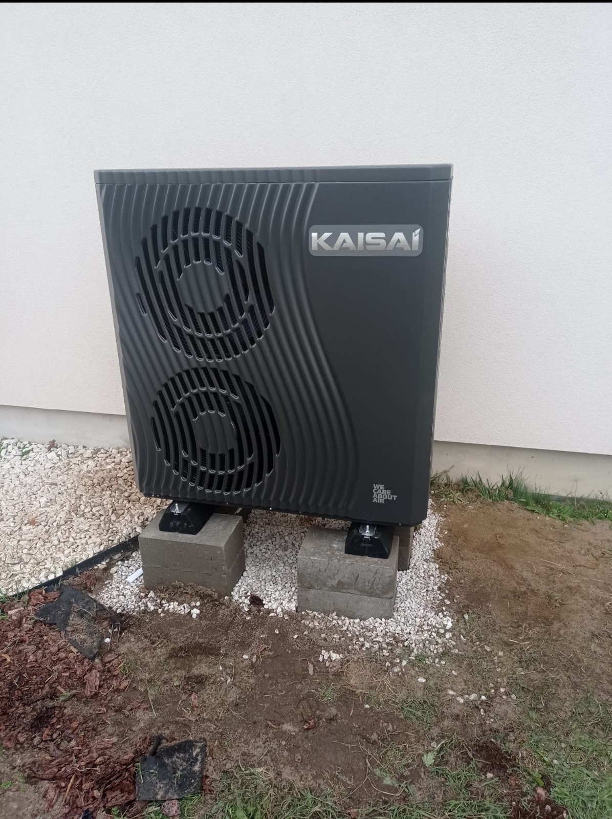 Czarna pompa ciepła Kaisai, umieszczona na betonowych bloczkach, z widocznym fragmentem elewacji budynku i otoczeniem z kamyków oraz ziemi.