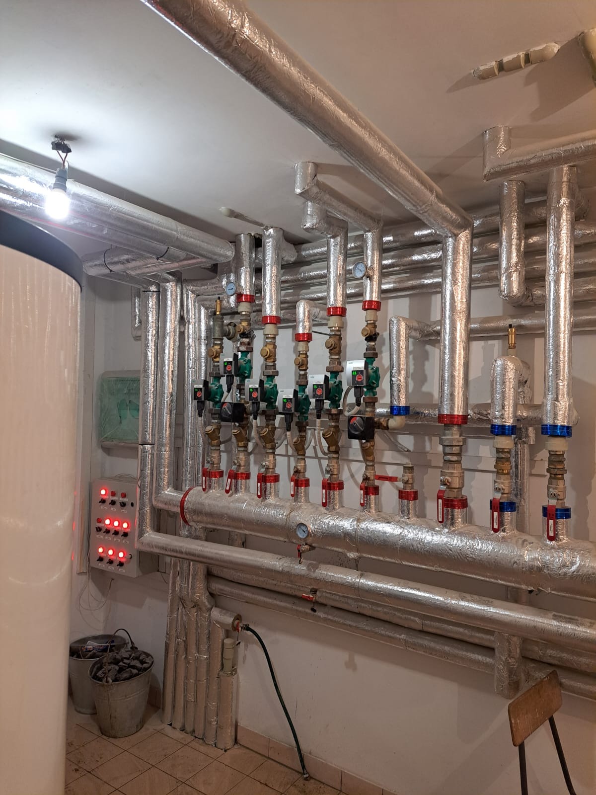 Rozbudowana instalacja grzewcza z rurami pokrytymi izolacją aluminiową, licznymi zaworami z czerwonymi i niebieskimi elementami, manometrami, oraz panelem sterowania z czerwonymi diodami, widoczne...