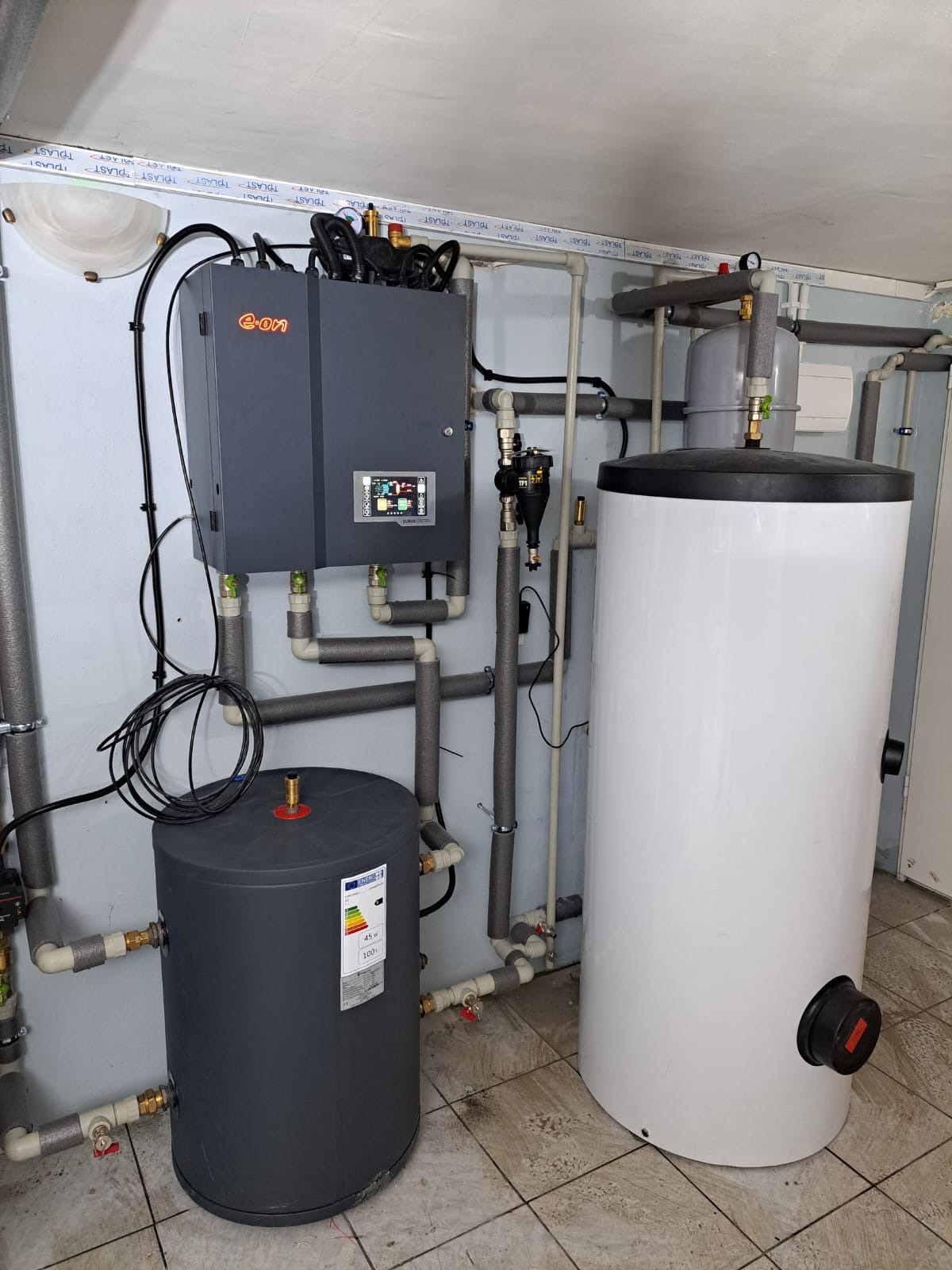 Zainstalowana instalacja grzewcza z szarym modułem sterującym E-on, dwoma zbiornikami na wodę w kolorze białym i szarym oraz rurami w izolacji termicznej na tle jasnej ściany i podłogi z płytek.