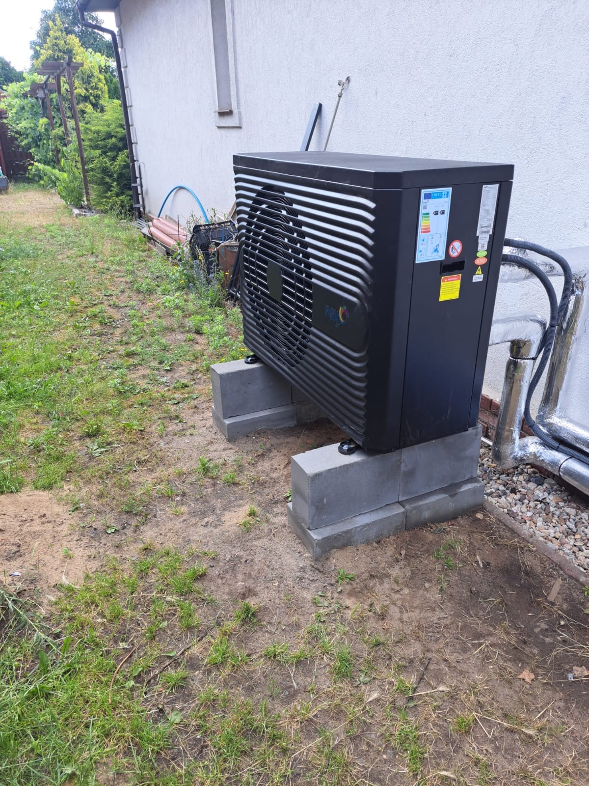 Czarna jednostka zewnętrzna urządzenia grzewczego, zainstalowana na betonowych bloczkach przy ścianie budynku, widoczne rury w izolacji.